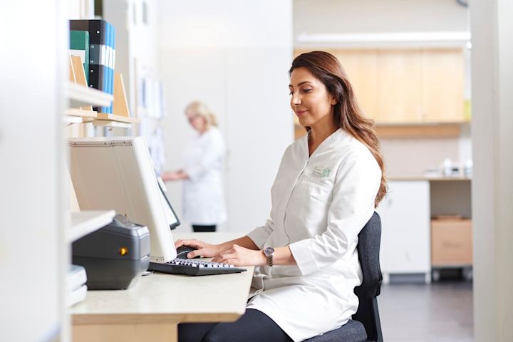 Kvinnlig vårdpersonal i vit rock sitter vid ett skrivbord och arbetar vid en dator på en klinik.