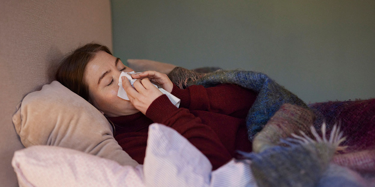 Förkyld kvinna ligger i sängen och snyter sig.