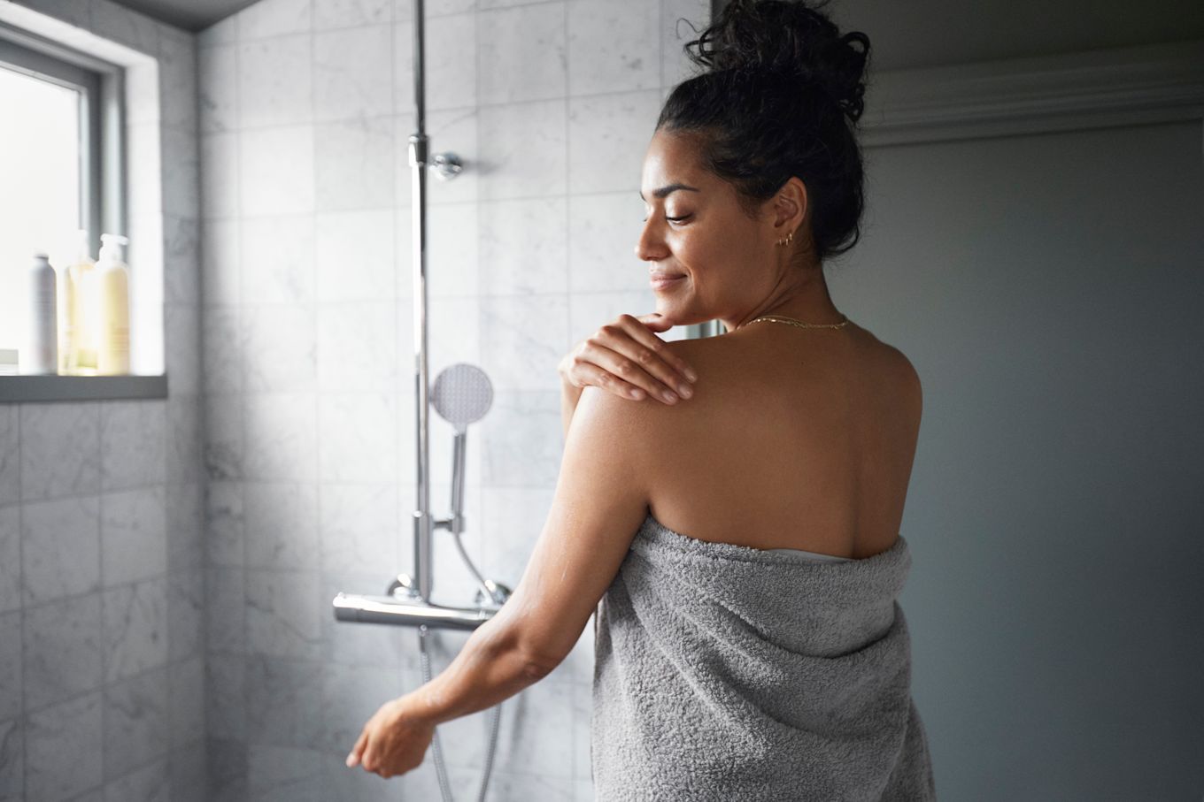 Kvinna står i badrum, invirad i handduk och smörjer in sin axel.