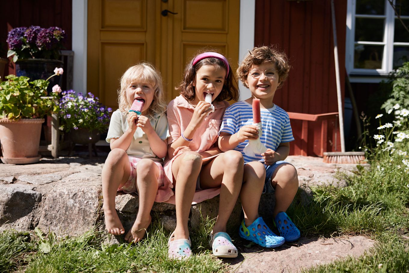 Tre barn sitter utomhus på sommaren i solskenet och äter glass tillsammans.