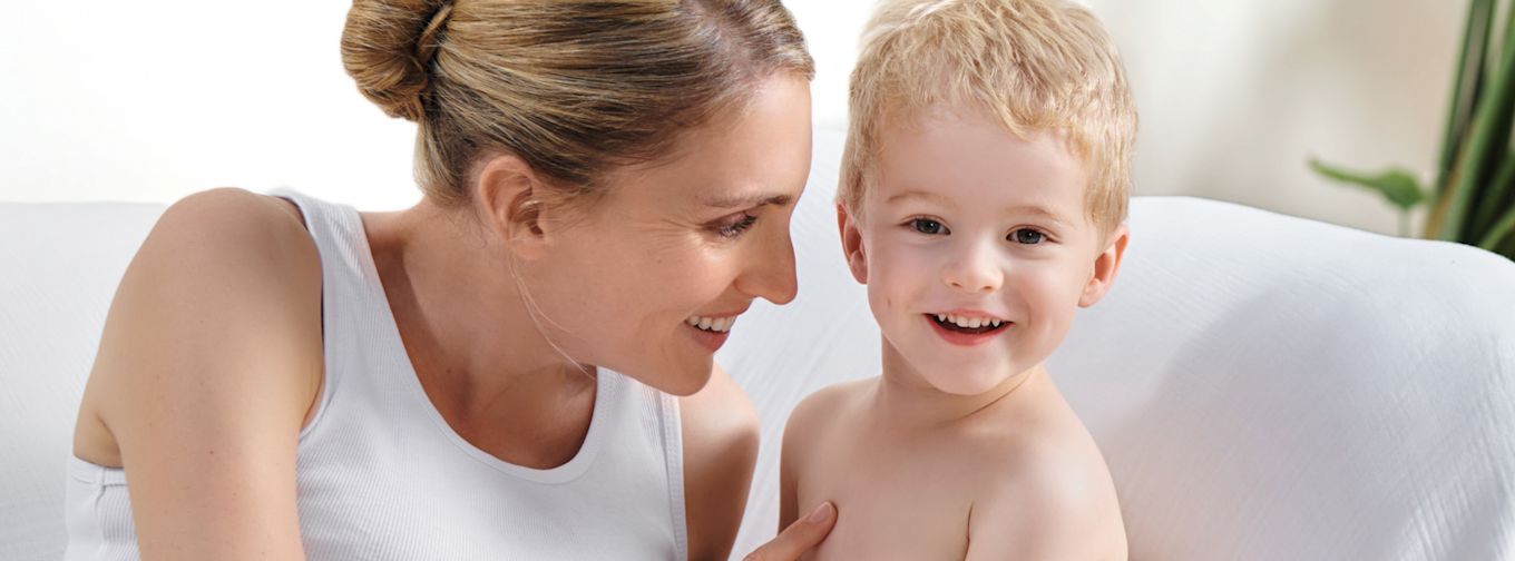 Leende kvinna sitter nära ett glatt litet barn på en vit soffa i ett ljust rum.