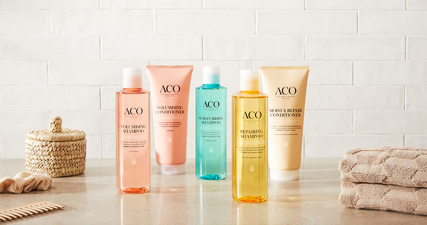 Olika produkter från ACO:s hårvårdsserie står uppradade på en bänk i badrummet. Bredvid ligger handdukar, en kam och ett hårband.
