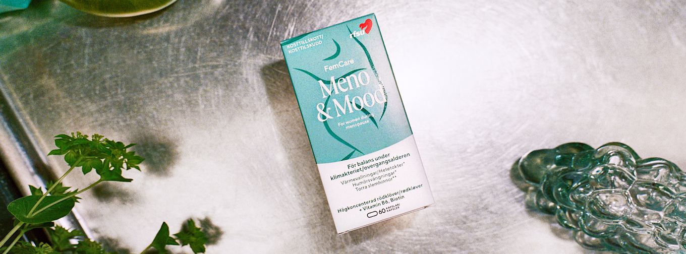 Förpackning av FemCare Meno & Mood kosttillskott ligger på en metallbänk, omgiven av gröna växter och ett glasföremål.