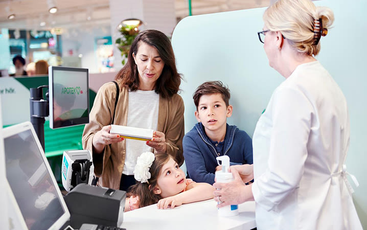 En familj står i kassan på ett apotek. Mamman läser på en förpackning och får råd av apotekaren medans barnen intresserat tittar på.