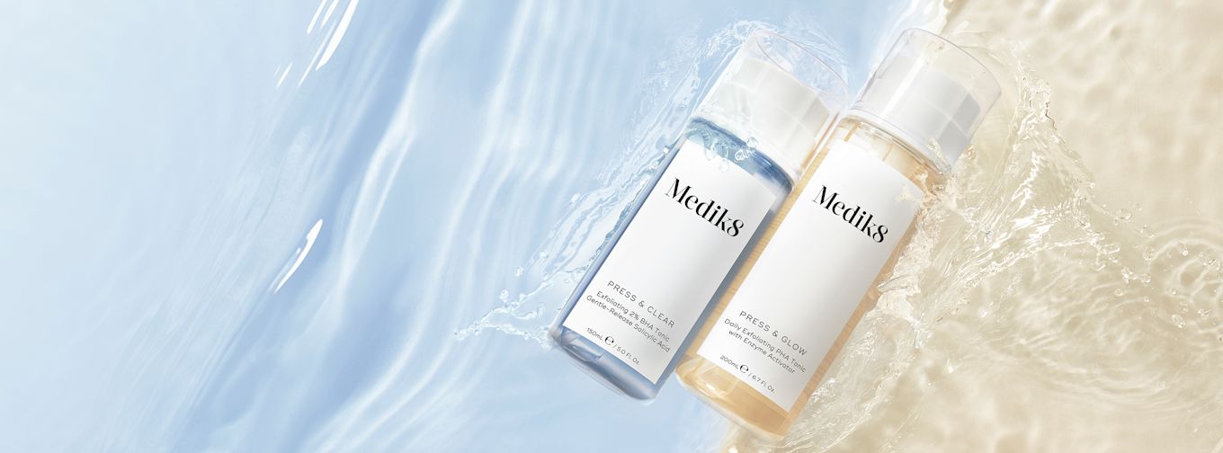 Två flaskor Medik8 ansiktsvatten, en blå och en gul, ligger på en våt yta med vattenstänk och ljus bakgrund.