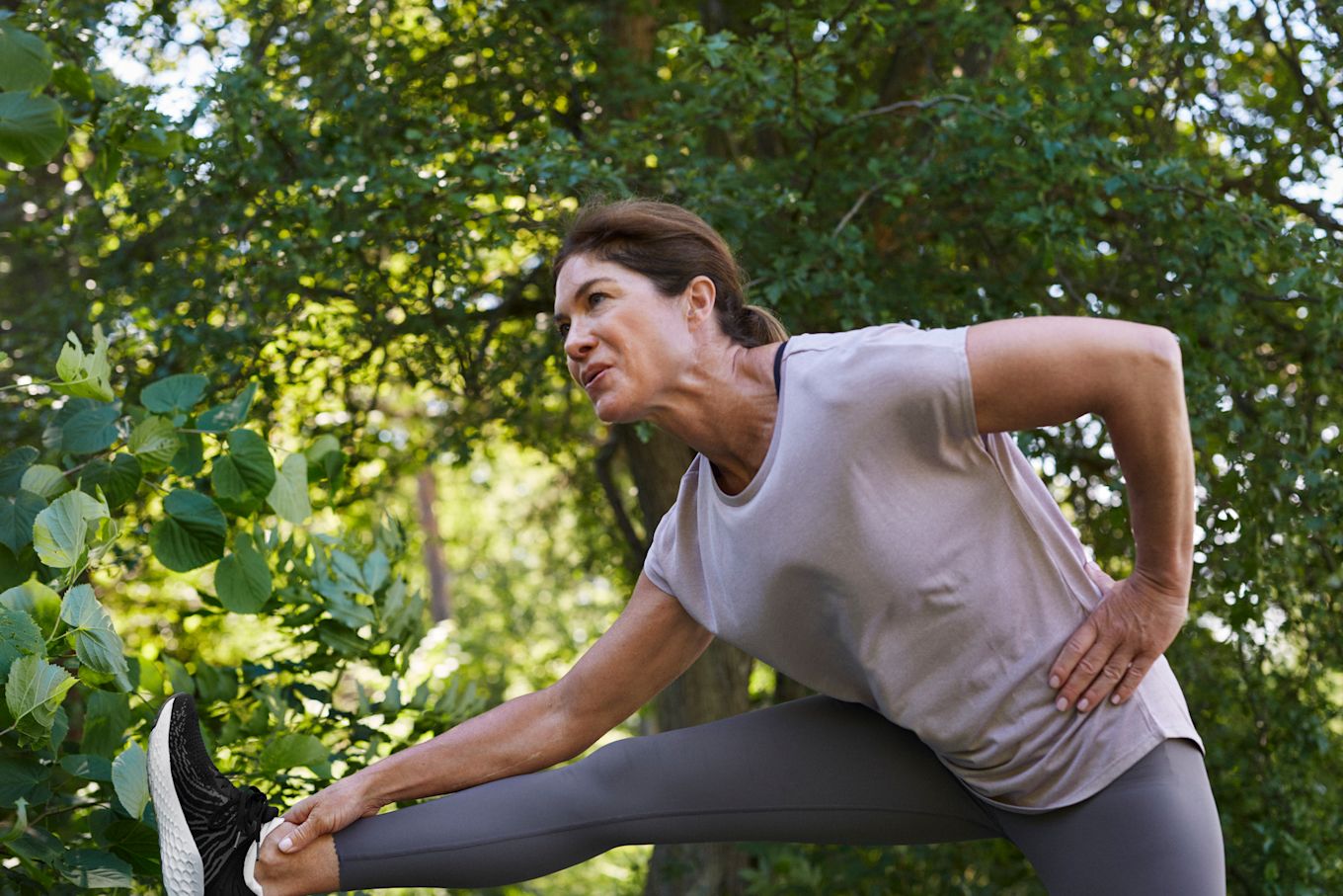 Kvinna står utomhus iklädd träningskläder med höger ben uppe och stretchar.