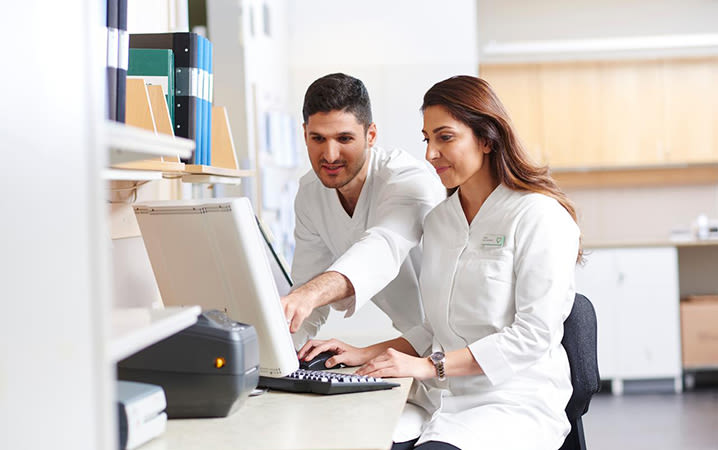 Två apotekare sitter inne på ett ljust kontor och samarbetar vid en dator.