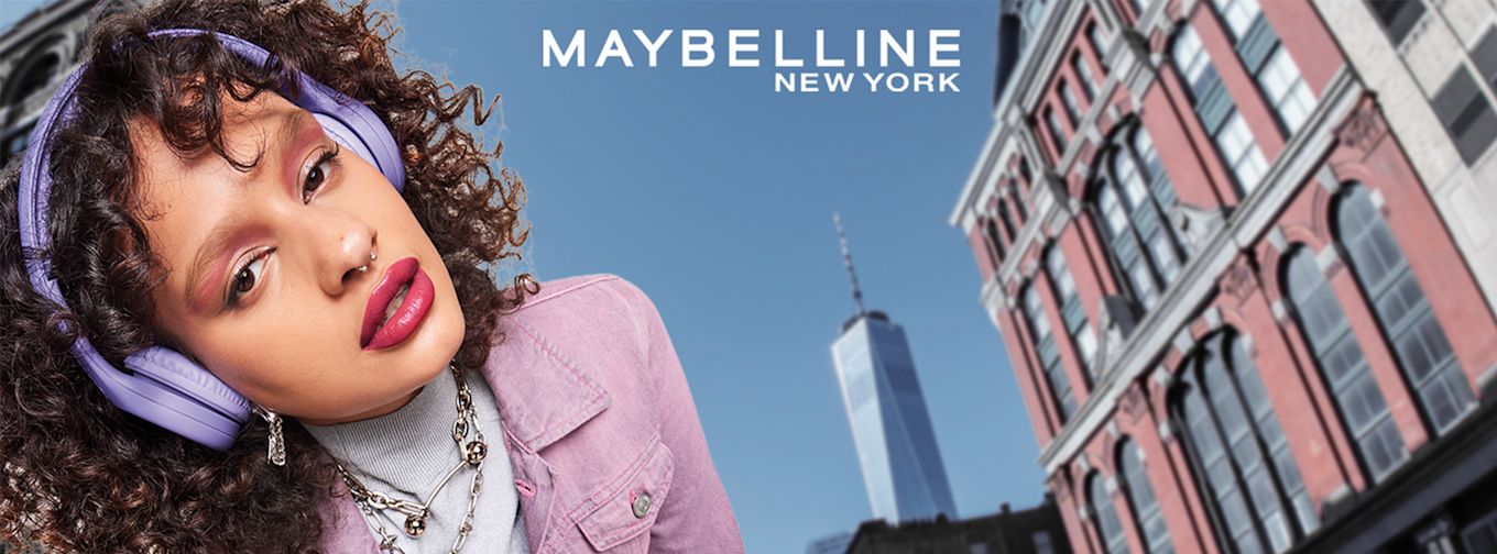 Kvinna med lockigt hår och lila hörlurar poserar i rosa jacka framför stadsmiljö, texten "Maybelline New York" syns ovanför.