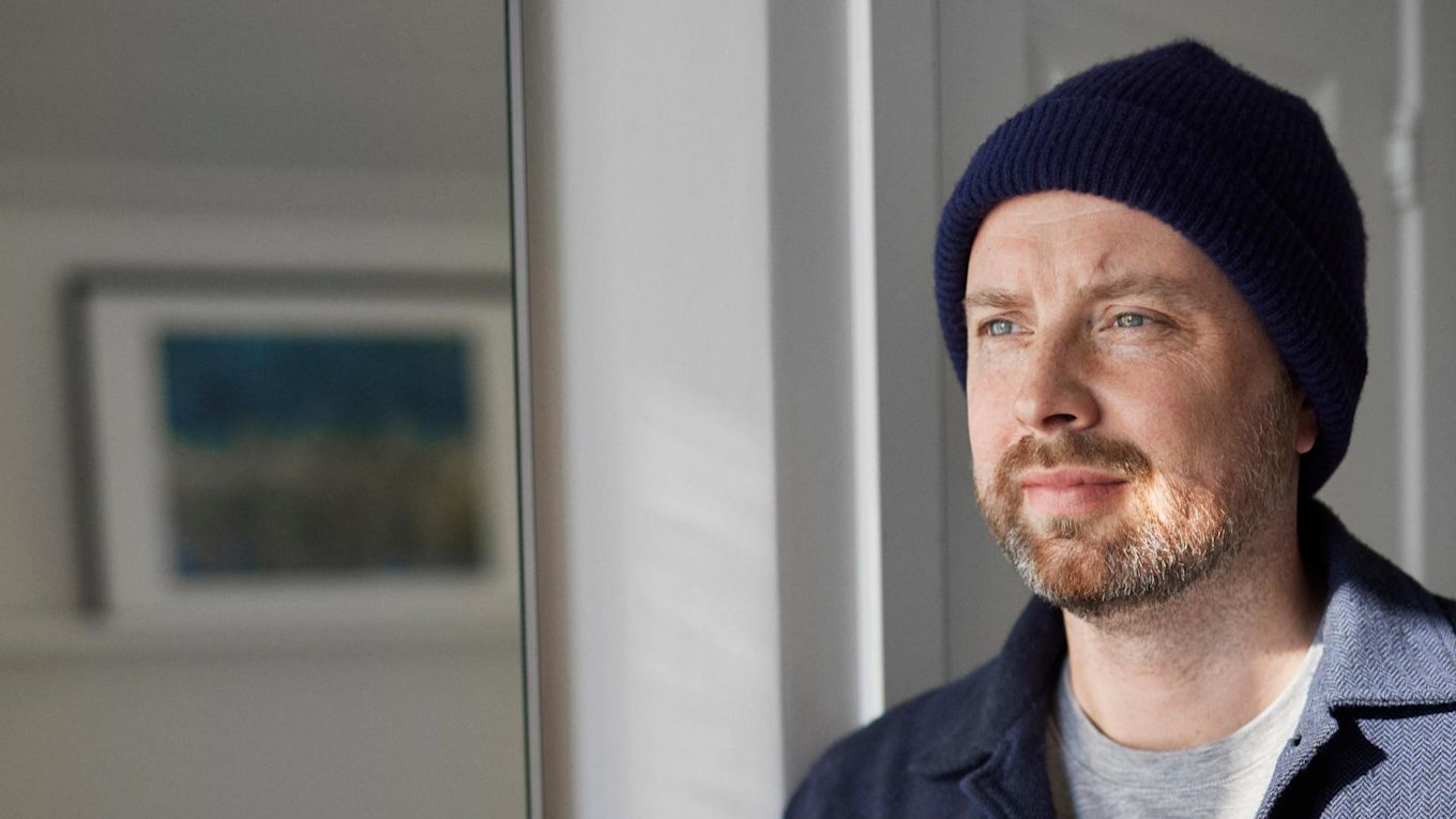 Man klädd i mörkblå jacka och mössa står vid ett fönster och tittar ut. 