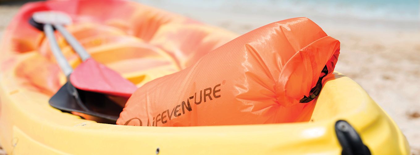 En orange vattentät påse med texten "HEVEN'URE" ligger i en gul kajak tillsammans med en paddel på en sandstrand.
