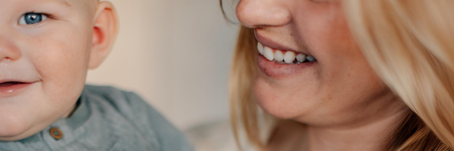 Närbild på en leende kvinna med blont hår som håller ett leende barn med blå ögon och grå tröja.
