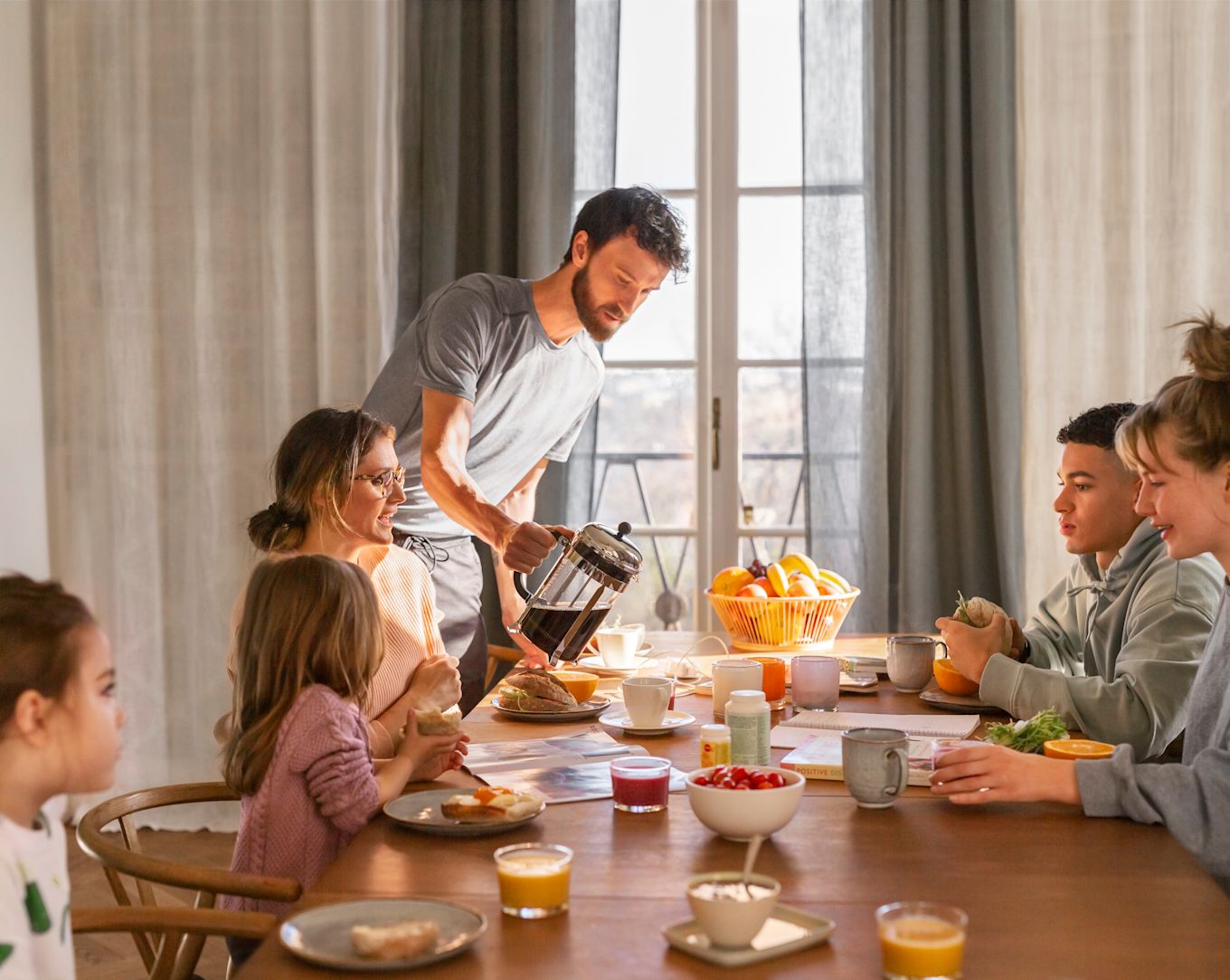 Familj sitter runt frukostbordet, fyllt med smörgåsar, juice, kaffe, frukt och yoghurt.