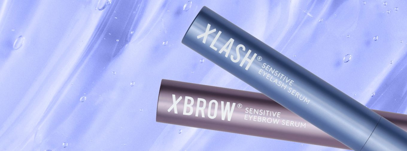 Två tuber med Xlash Sensitive Eyelash Serum och Xbrow Sensitive Eyebrow Serum ligger mot en ljuslila bakgrund med vattendroppar.