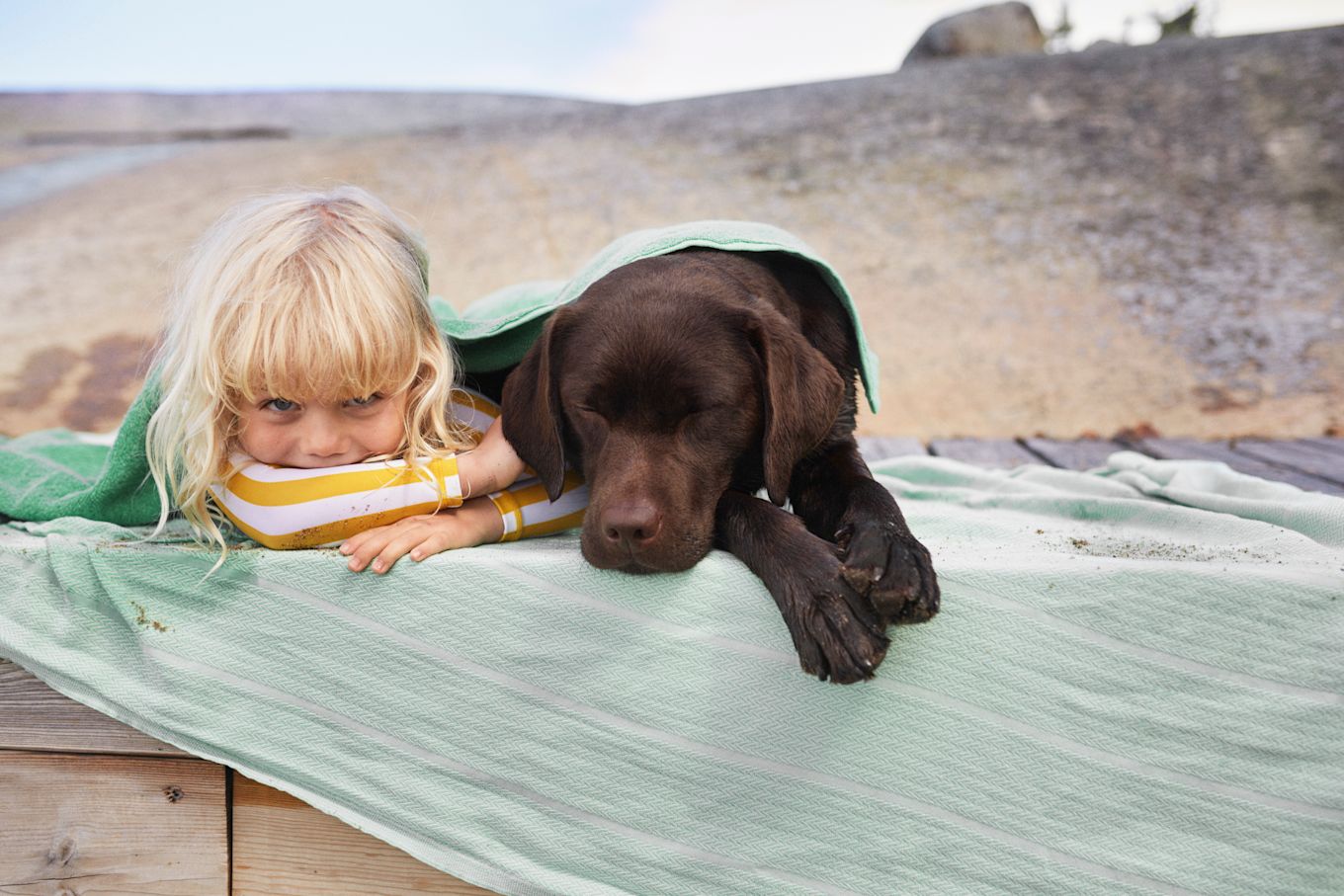 Flicka och hund ligger och vilar under en filt på stranden.