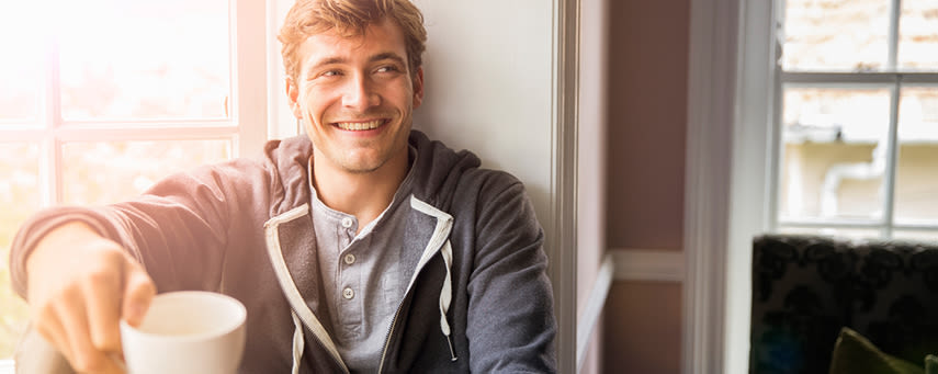 Kille sitter på fönsterbrädan och ler. Han håller en kaffekopp i handen.