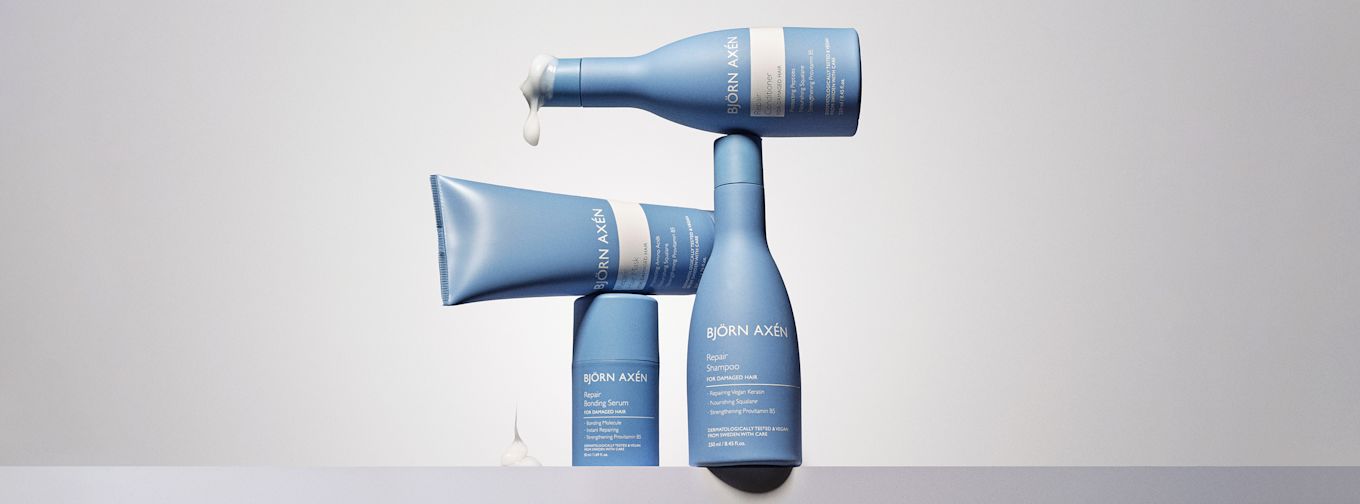 Tre blå Björn Axén hårvårdsprodukter står staplade, med schampo, balsam och inpackning, mot ljus bakgrund. Kräm rinner från balsamflaskan.