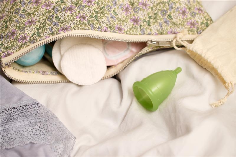 En blommig påse som delvis öppnas upp, avslöjar bomullsrondeller och en grön menskopp på ett vitt överkast.