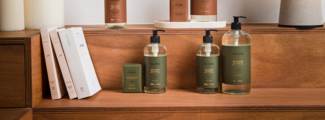 Tre flaskor och en tvål från Fer à Cheval står på en trähylla bredvid böcker och dekorativa föremål.