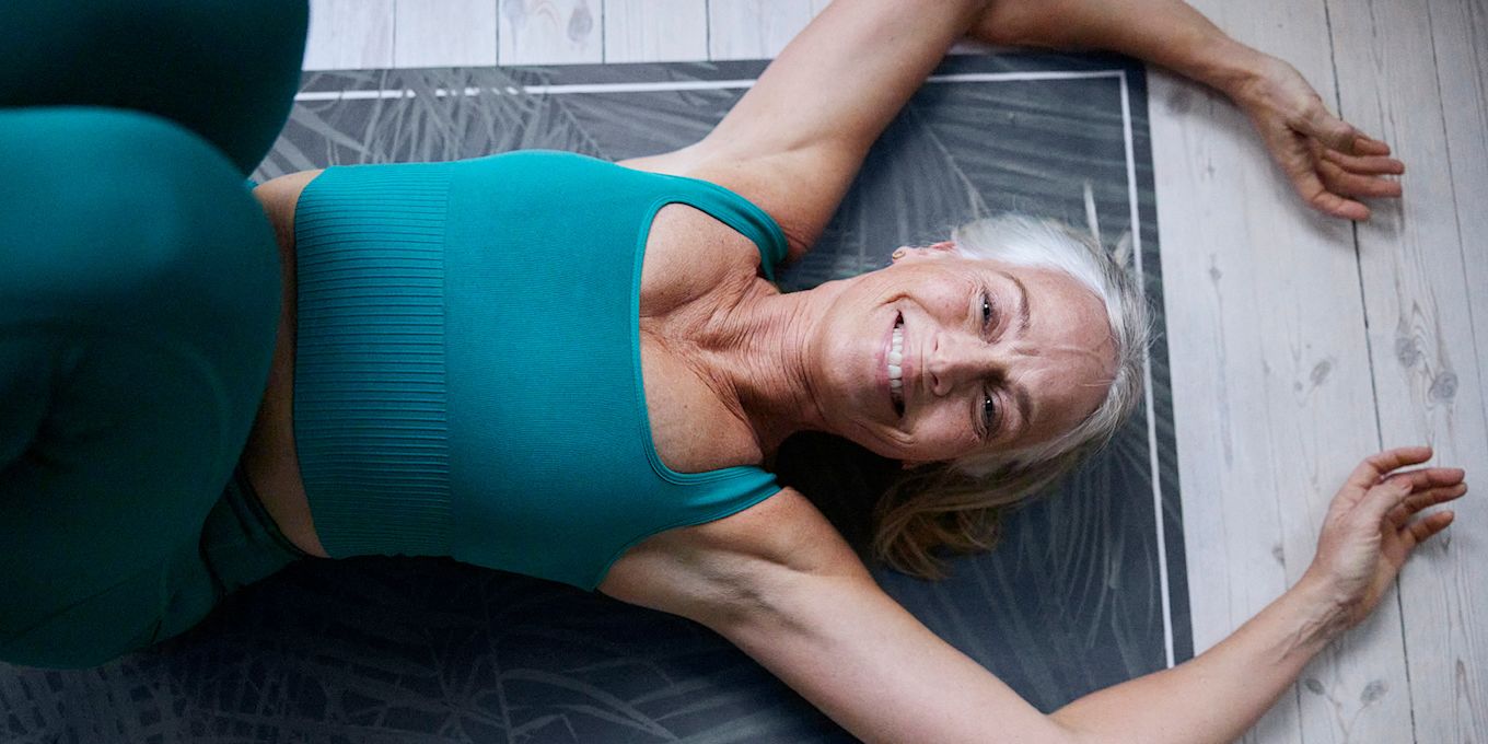 En äldre kvinna ligger på en yogamatta på golvet, med armarna över huvudet och ler.
