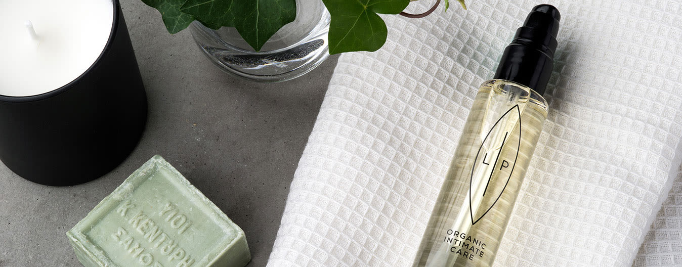 Flaska med LP Organic Intimate Care-olja ligger på en vit handduk bredvid ett ljus, grön tvål och en glasvas med murgröna.
