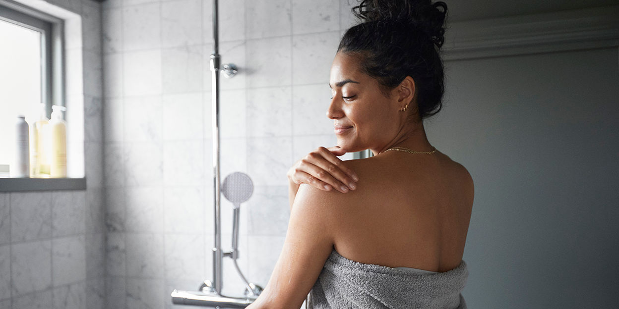 Kvinna står med ryggen mot kameran i badrummet med en handduk runtomkring sig. Hon kollar snett bakåt. 