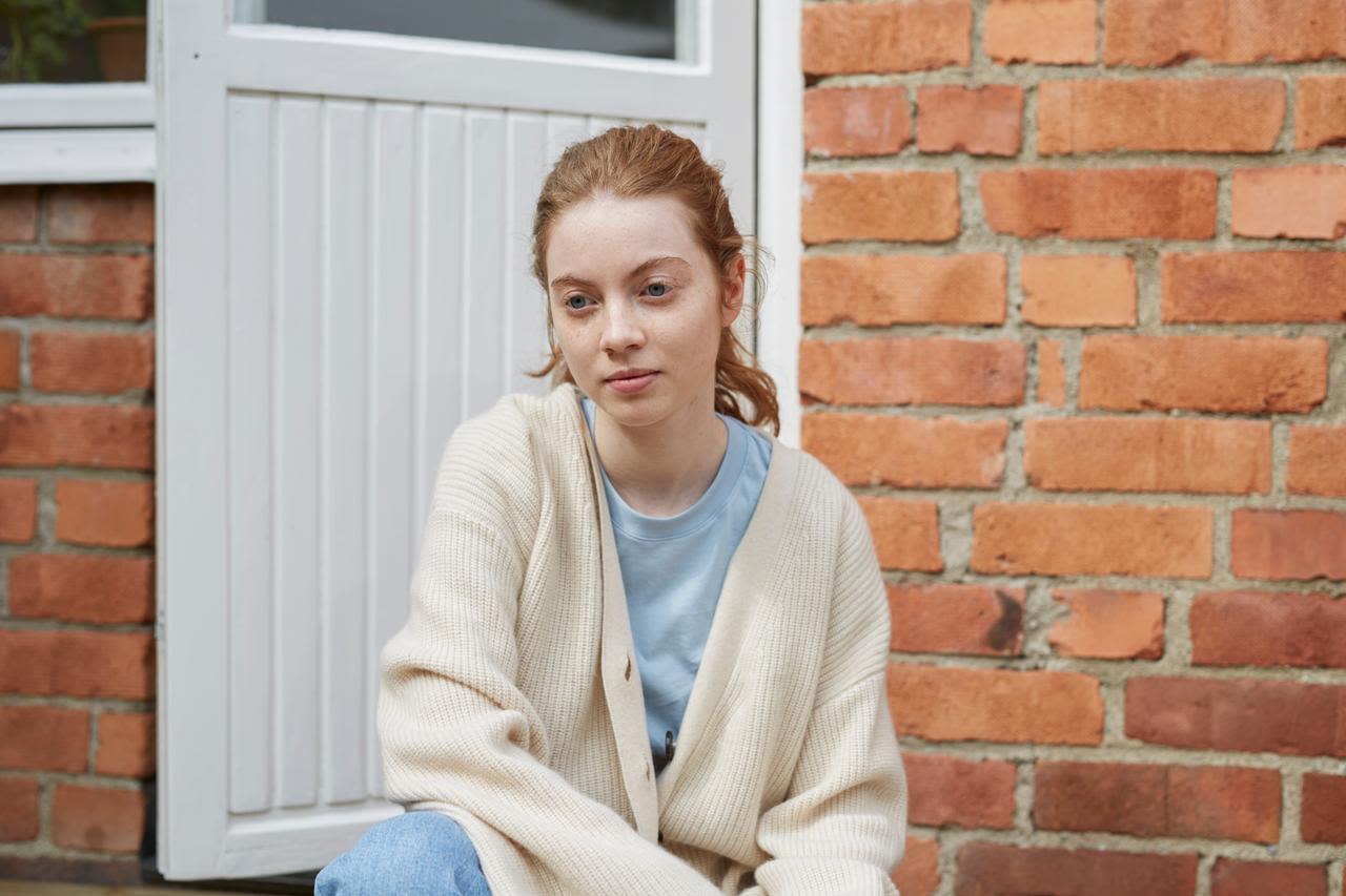 Rödhårig, ljus ung kvinna sitter utanför ett rött tegelhus med vit dörr. hon har en ljusblå t-shirt och en vit stickad kofta på sig. Ansiktsuttrycket är neutralt. 