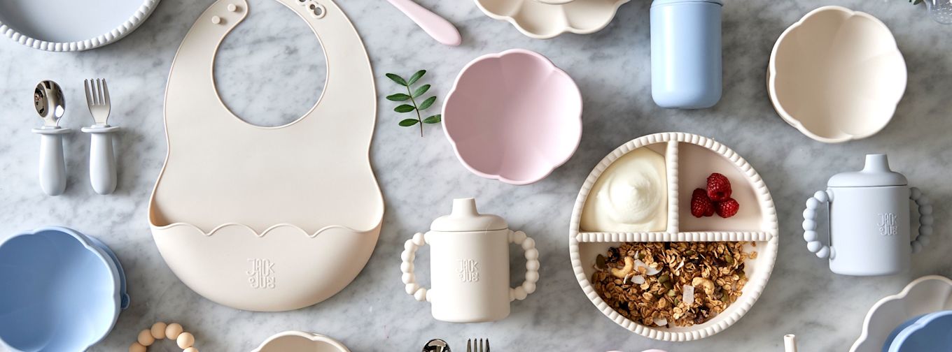 Barnservis i pastellfärger med tallrik, skål, haklapp, pipmugg, bestick och delad tallrik med yoghurt, granola och hallon på marmorbord.