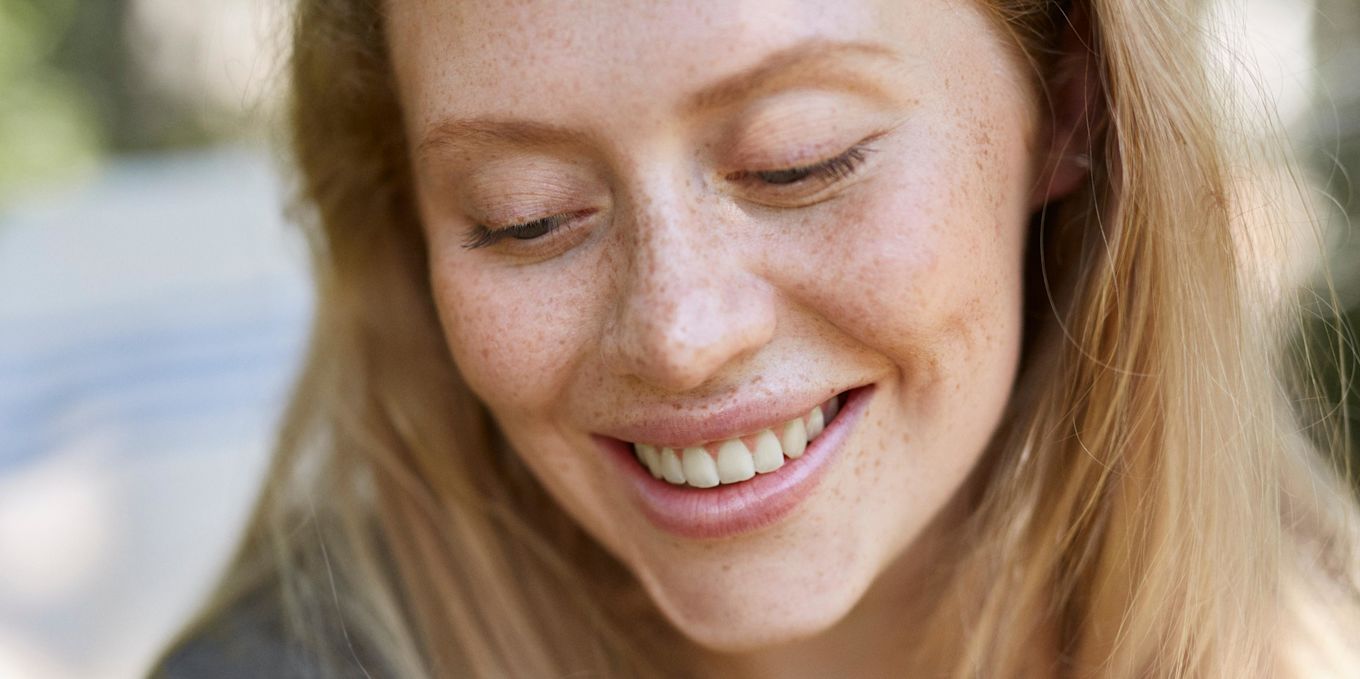 Närbild på en leende person med fräknar som tittar ner.