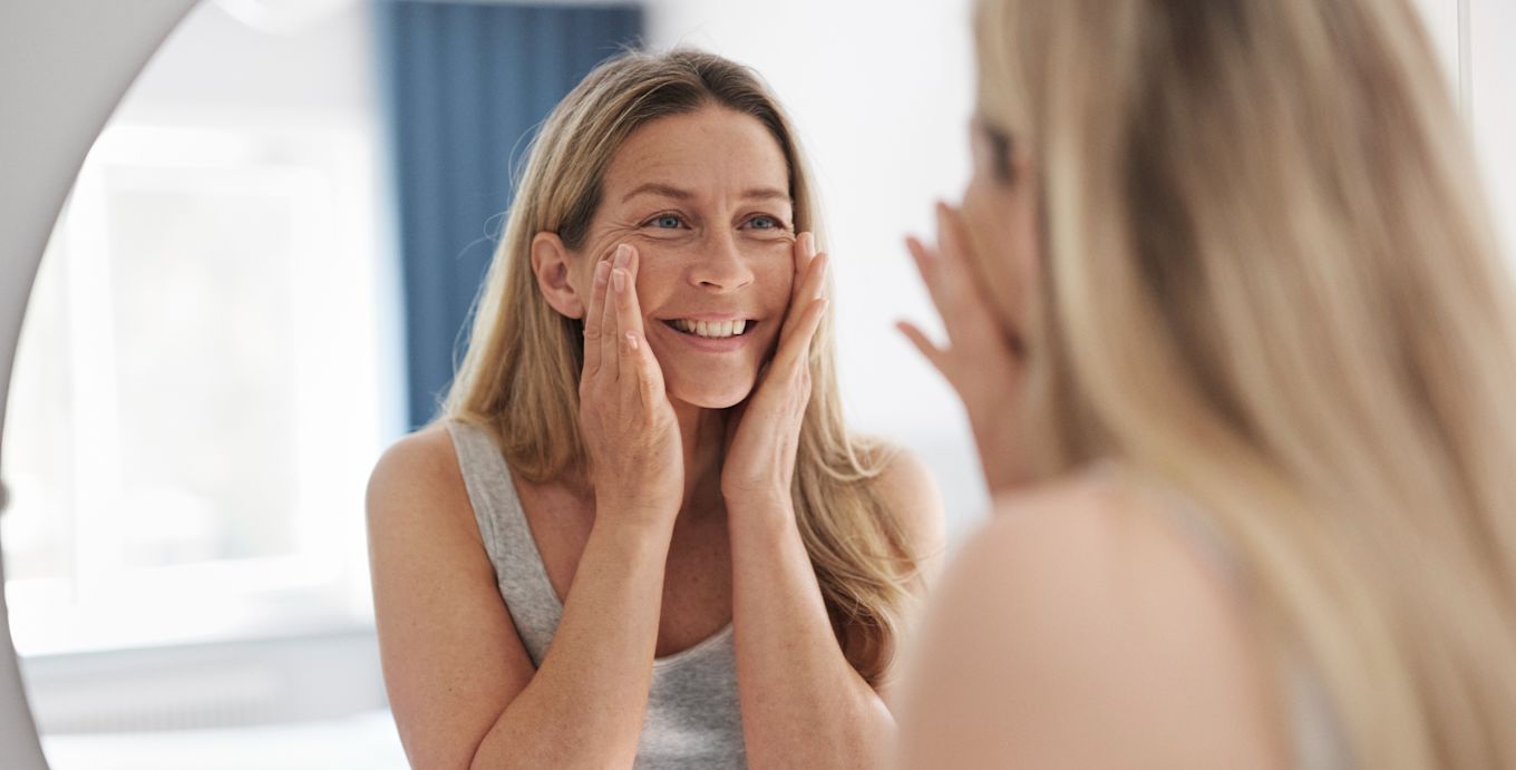Kvinna tittar på sin spegelbild, rör vid sitt ansikte med sina händer och ler.