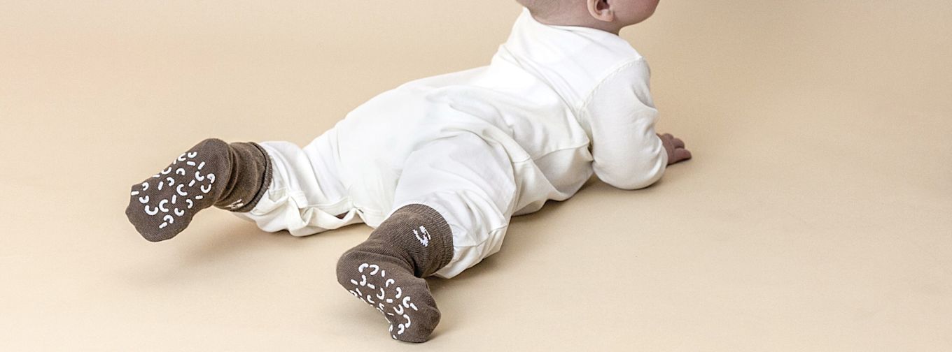 Bebis i vit sparkdräkt ligger på mage på beige underlag och bär bruna halksockor med vita mönster.