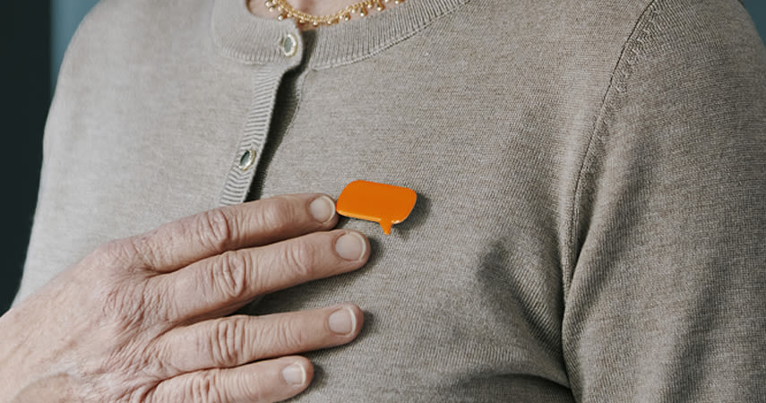 En person bär en beige kofta med en orange pratbubblemärke fäst vid bröstet och håller handen över märket.