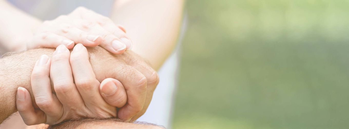 Två händer håller varsamt om en annan hand som en gest av stöd och omtanke, med suddig grön bakgrund.