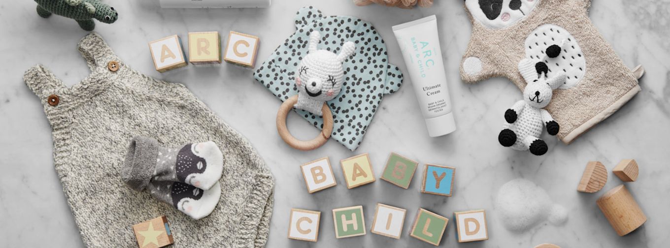 Stickad barnklänning, babysockor, träklossar med texten "ARC BABY CHILD", babykräm, gosedjur och leksaker på marmorbord.
