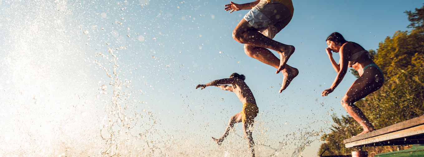 Tre personer hoppar från en brygga ner i vattnet, vattenstänk fyller luften, soligt väder och träd i bakgrunden.