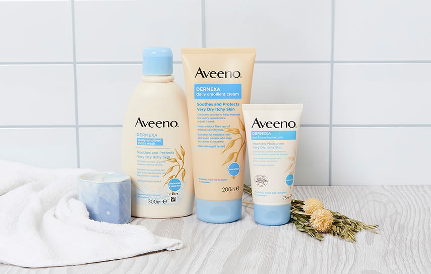 I ett badrum står tre produkter från Aveeno uppradade. Bredvid ligger en vit handduk och gula blommor.