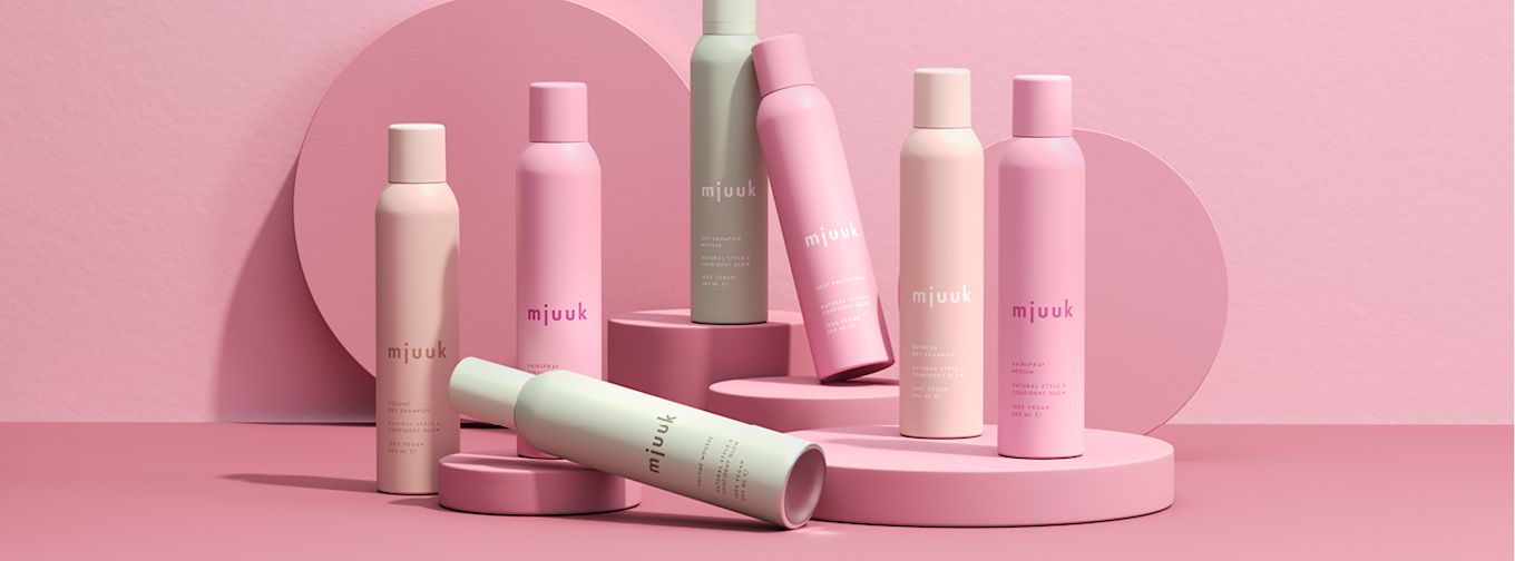 Flera pastellfärgade flaskor med texten "mjuuk" står och ligger på rosa podier mot en rosa bakgrund. Produkterna är hårvård.