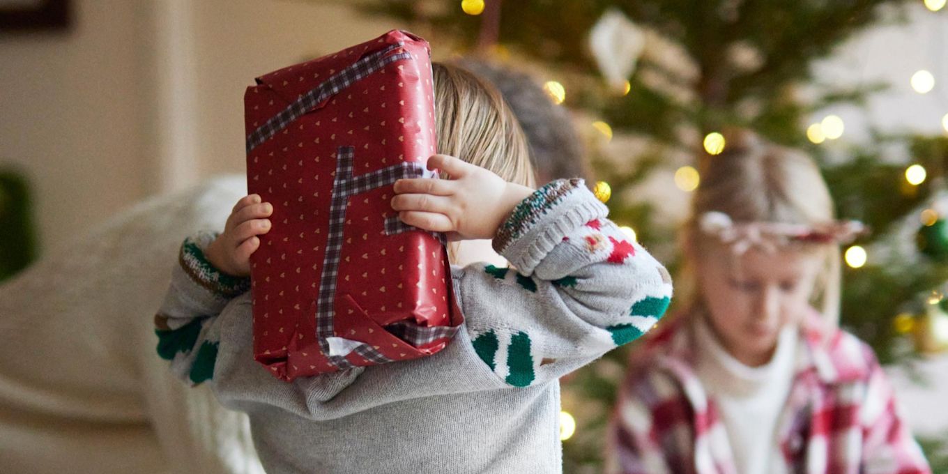 Ett litet barn står med en inslagen julklapp bakom ryggen. Framför står en pyntad julgran, och ett annat barn öppnar ett paket.