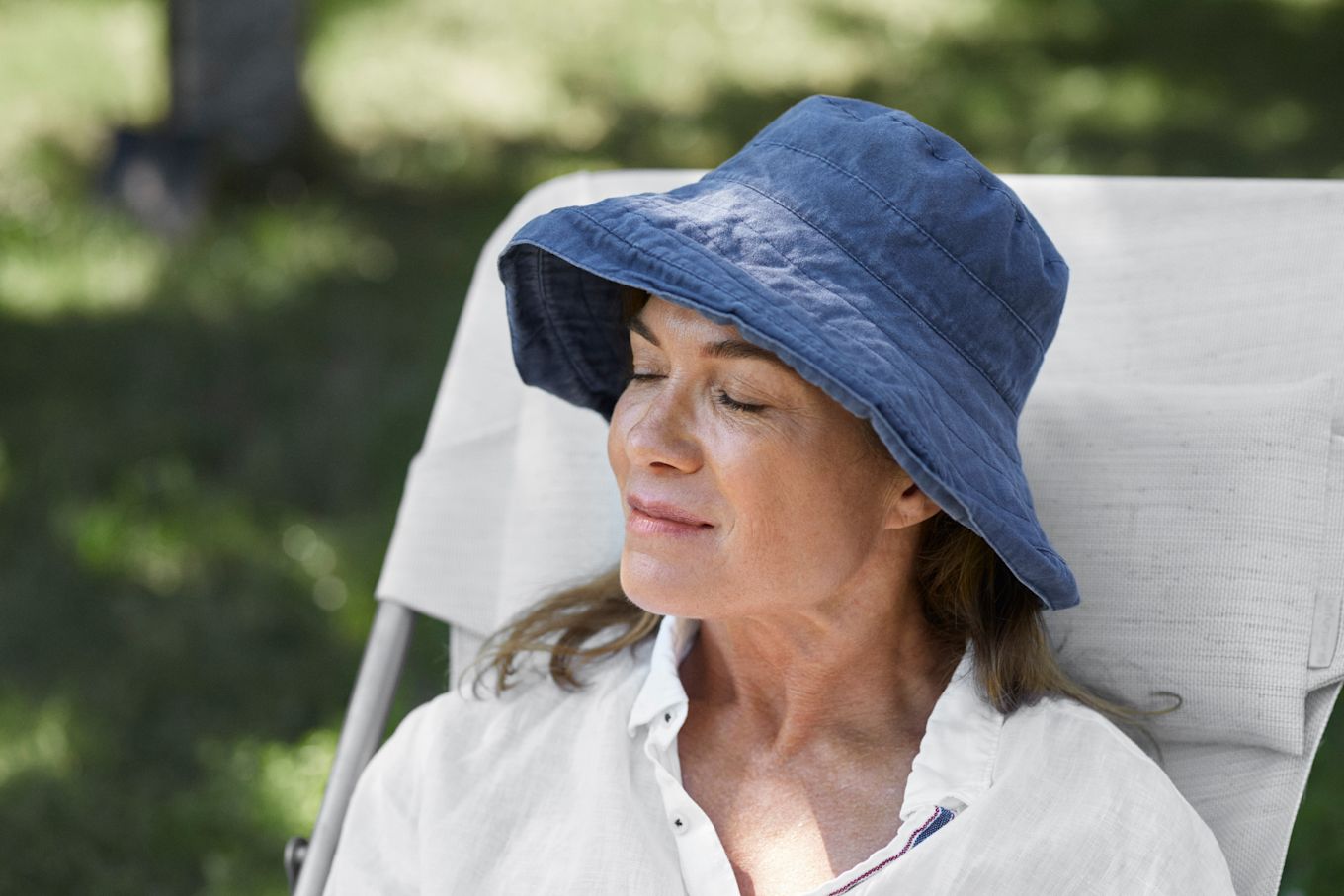 Kvinna sitter utomhus i en solstol och blundar, med en blå solhatt.