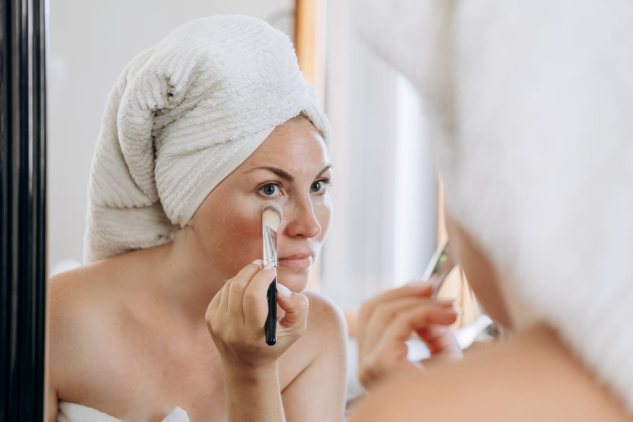 Kvinna står framför spegel och applicerar smink med en borste, hon har en handduk virad runt kroppen och håret uppsatt med en handduk. 