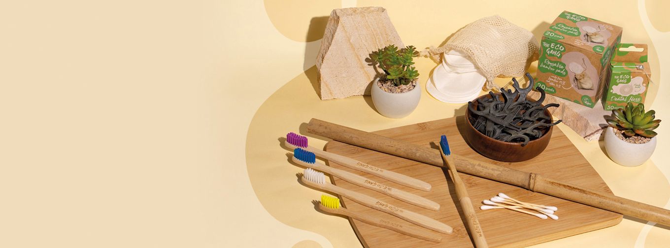 Fyra bambutandborstar med färgade borst, tandtråd, bomullspinnar, återanvändbara bomullsrondeller och små krukväxter på en träbricka.