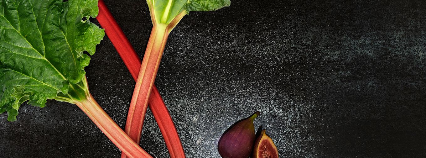Två rabarberstjälkar med gröna blad ligger bredvid ett helt och ett halvt fikon på en mörk yta.