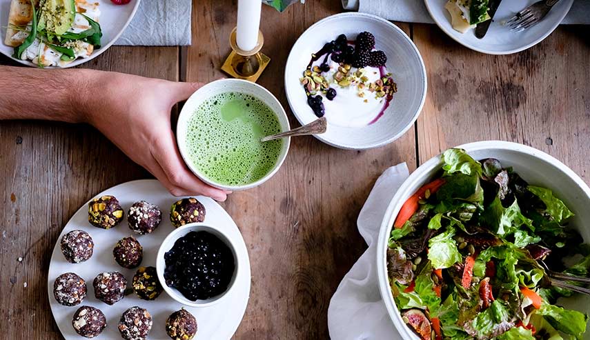 På ett matbord står en blanding av tallrikar och skålar med bland annat sallad, grön soppa och yoghurt med bär. En hand sträcker sig in och tar tag i skålen med grön soppa.
