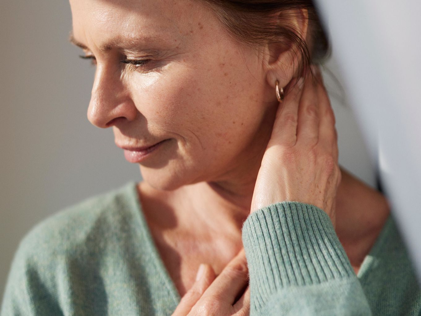 Kvinna står i solen och tittar åt sidan. Hon håller upp ena handen mot sin nacke.