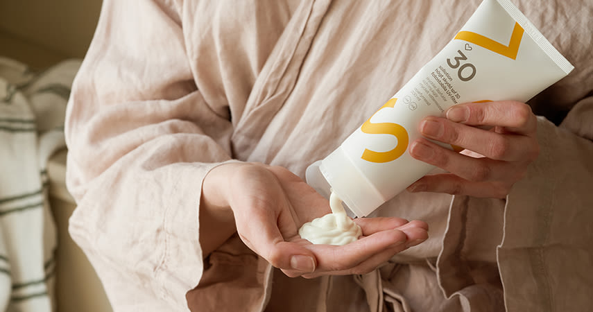 Person trycker ut solkräm med SPF 30 från en tub i handen, iförd beige morgonrock.
