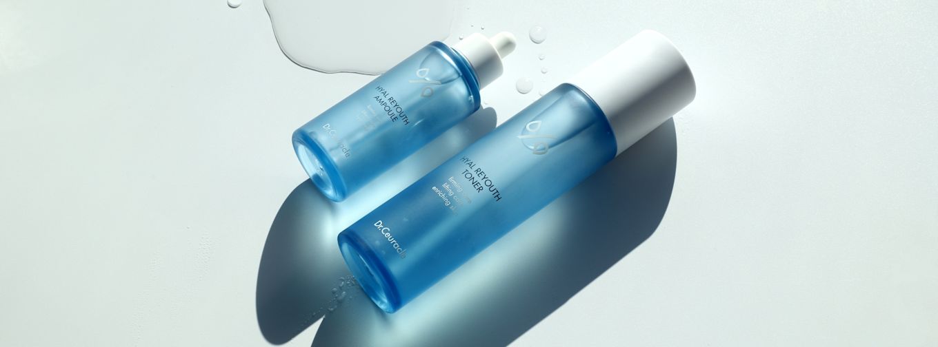 Två blå flaskor med vita lock från Dr.Ceuracle, märkta "HYAL REYOUTH AMPOULE" och "HYAL REYOUTH TONER", ligger på ett vitt underlag med vattendroppar.