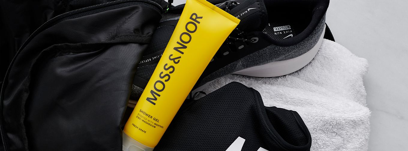 Gul tub MOSS & NOOR duschgel ligger på en vit handduk bredvid svarta träningsskor, väska och svart tröja.