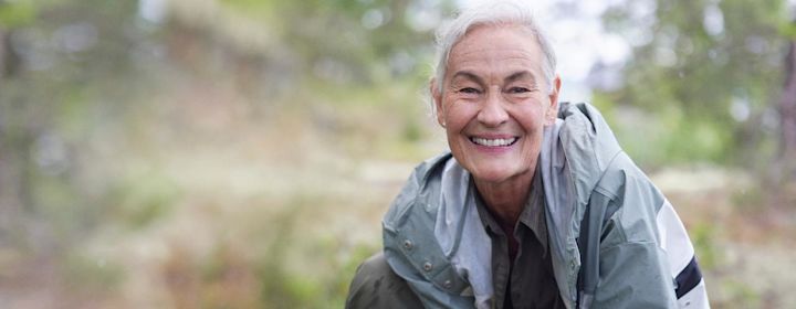 Glad äldre kvinna ler stort i skogen iklädd en regnjacka.