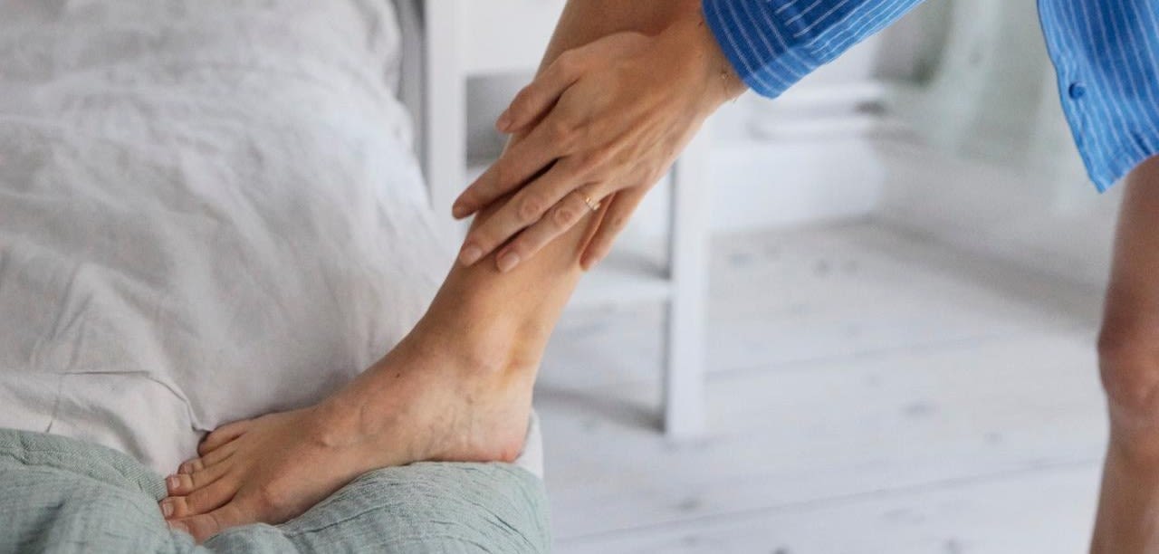 En kvinna med bara ben i blå- och vit-randig skjorta som har satt upp sjn fot på sängkanten. Hon stryker sin hand mot sitt bara ben från fot upp mot knä.