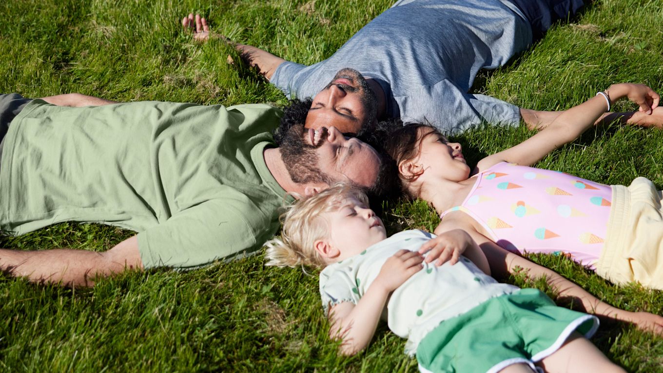 Två män och två barn ligger utomhus på en gräsmatta i solen.